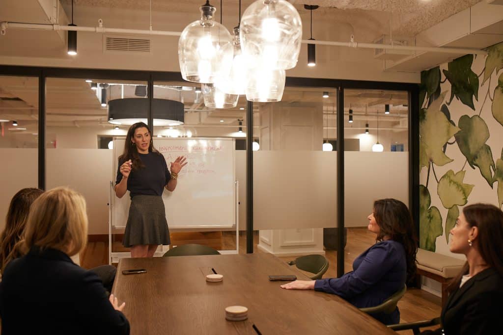 A woman stands and presents in front of a whiteboard to three seated women in a modern conference room with glass walls, pendant lights, and leafy wall decor—an inspiring setting for NYC Recruiters.