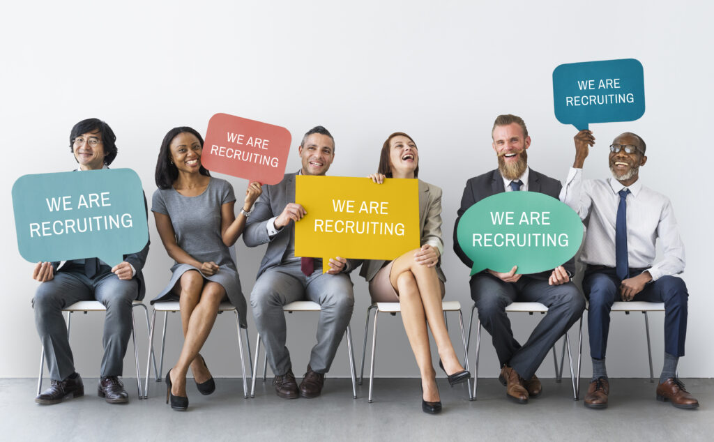 Six professionally dressed people sit on chairs, smiling and holding colorful signs that say “WE ARE RECRUITING,” highlighting the spirit of Competing for Leadership Talent in a Tight Market against a plain white background.