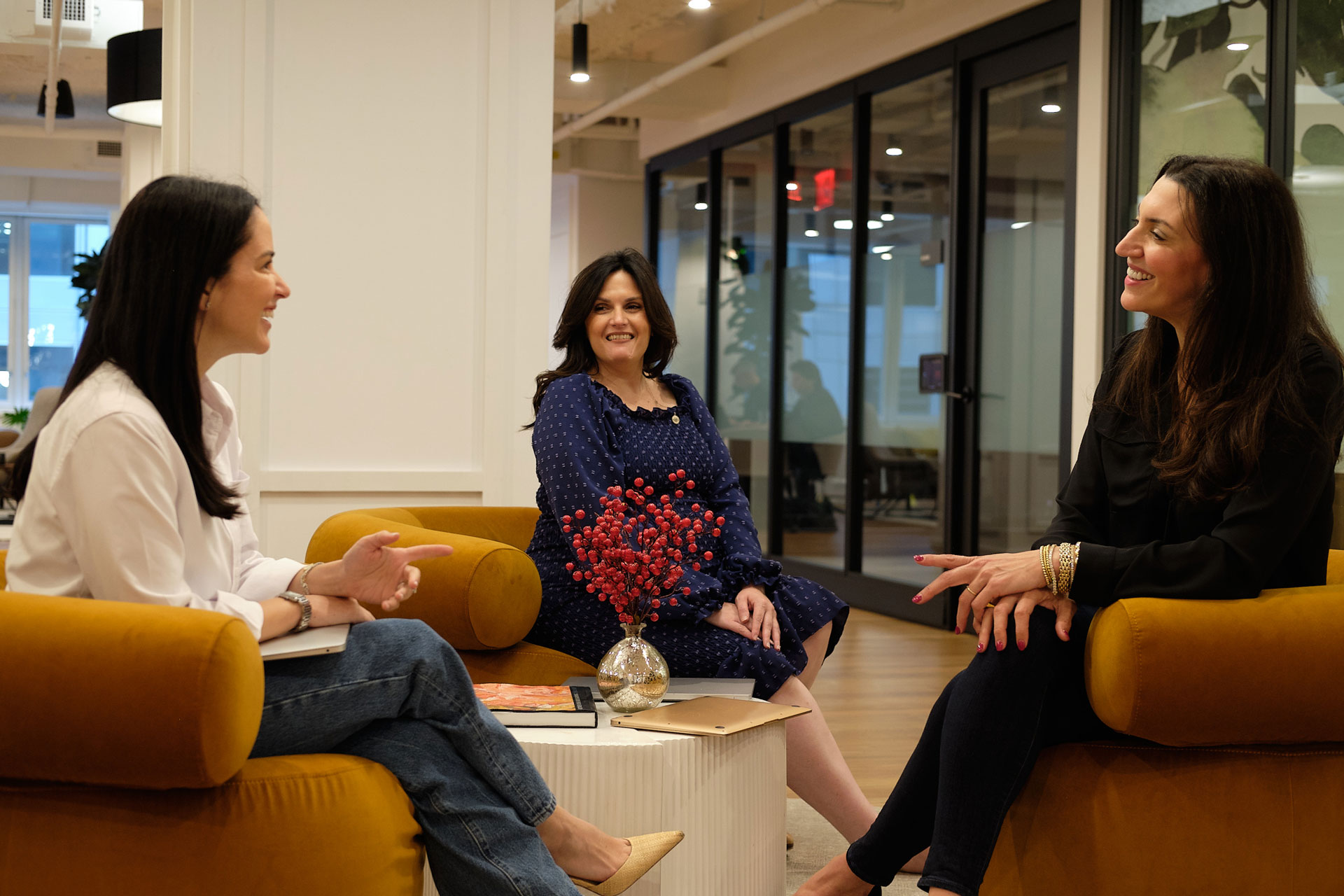 Three women sit together on mustard-colored chairs around a small table, smiling and talking in a modern creative media office space. A small vase with red berries is on the table in front of them.