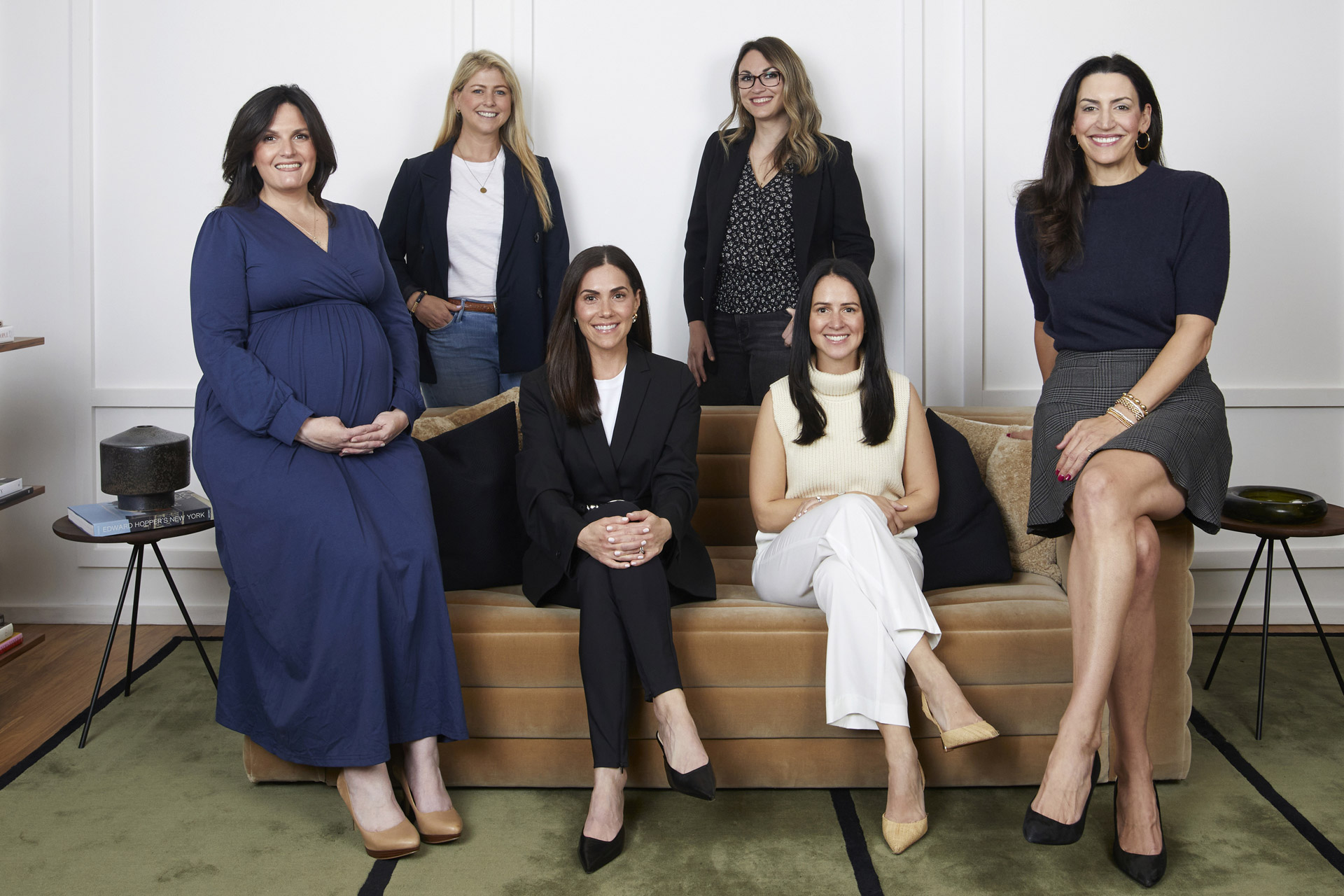 Six women pose together, three sitting and three standing behind a tan sofa, all smiling. Dressed in chic business-casual outfits, they exude professionalism in a modern living room—reflecting the dynamic team at an Executive Assistant Placement Agency in NYC.