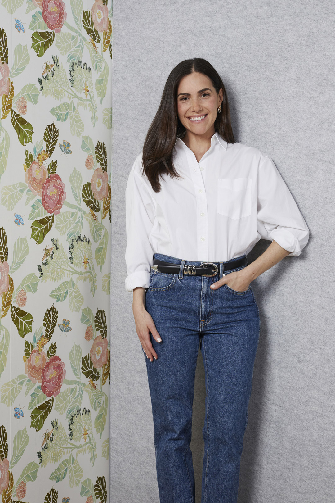 A woman with long dark hair, wearing a white button-up shirt and blue jeans with a black belt, stands smiling beside a wall decorated with a colorful floral pattern.