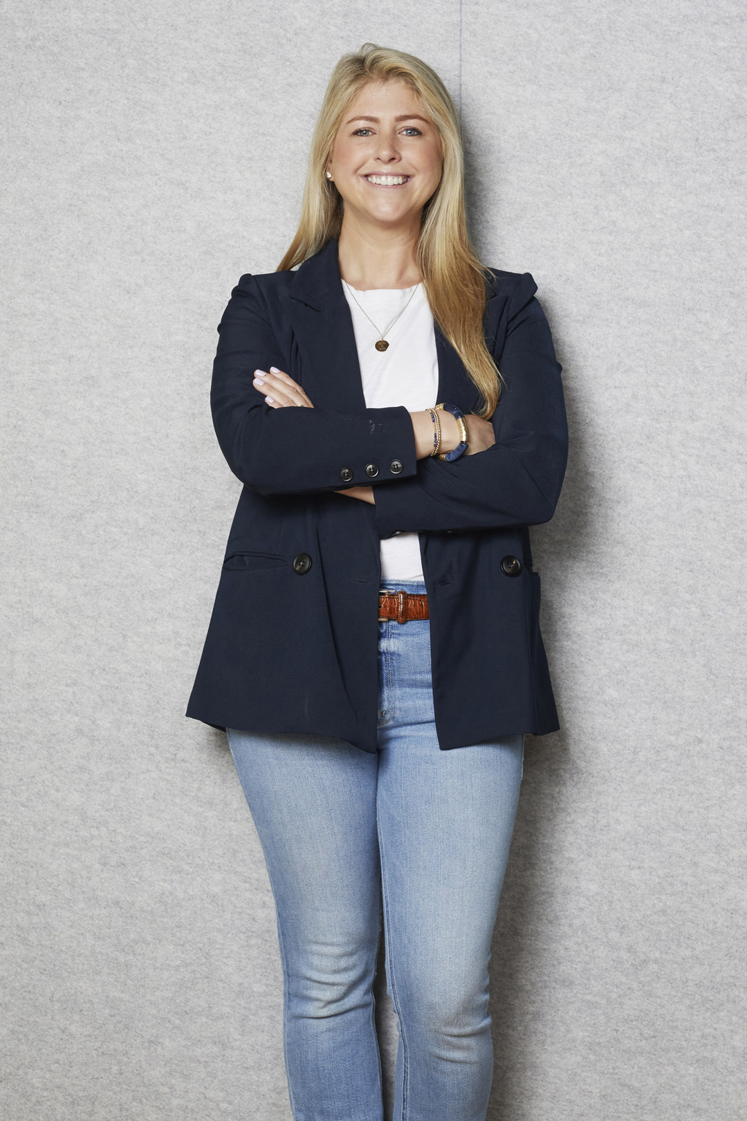 A woman with long blonde hair, wearing a navy blazer, white shirt, blue jeans, and brown belt, stands smiling with arms crossed in front of a light gray background.