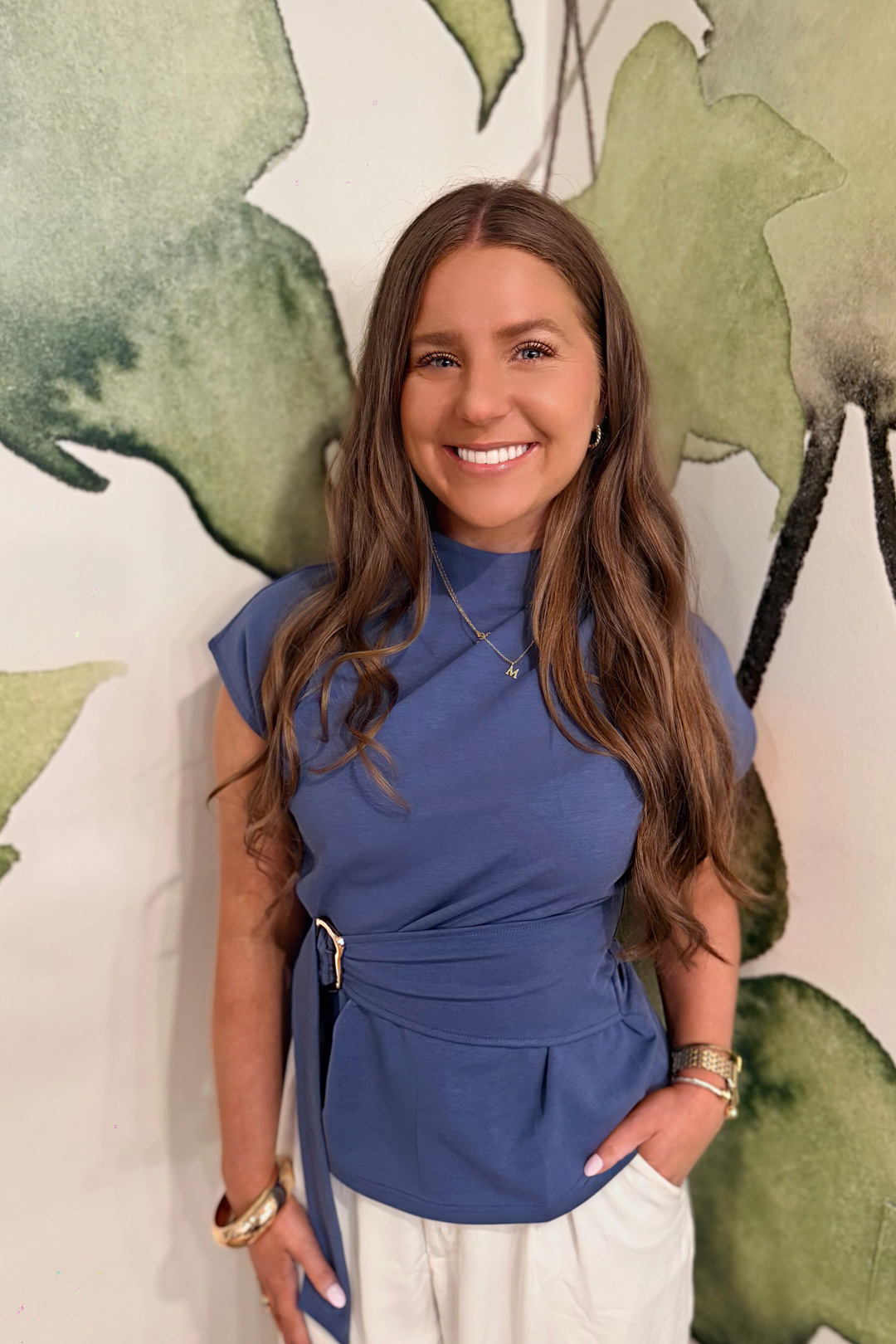 A woman with long brown hair wearing a blue top and white pants stands smiling in front of a wall with a green and white abstract leaf design. She has one hand in her pocket and wears jewelry on both wrists.