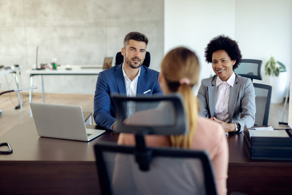 Two professionals, a man and a woman, sit at a desk with a laptop, interviewing a woman in a modern office setting, illustrating how search firms vet candidates beyond the resume.