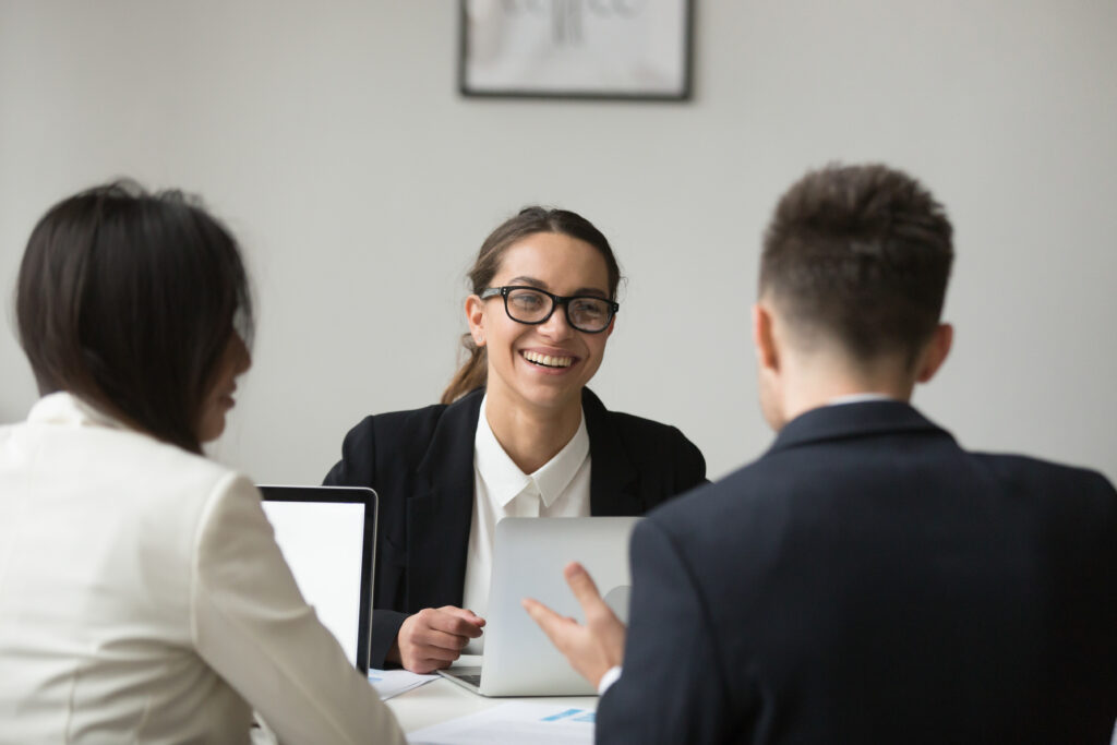 Three people in business attire sit at a table with laptops, discussing how search firms vet candidates beyond the resume. A woman in glasses smiles at two colleagues, creating a friendly and professional atmosphere.