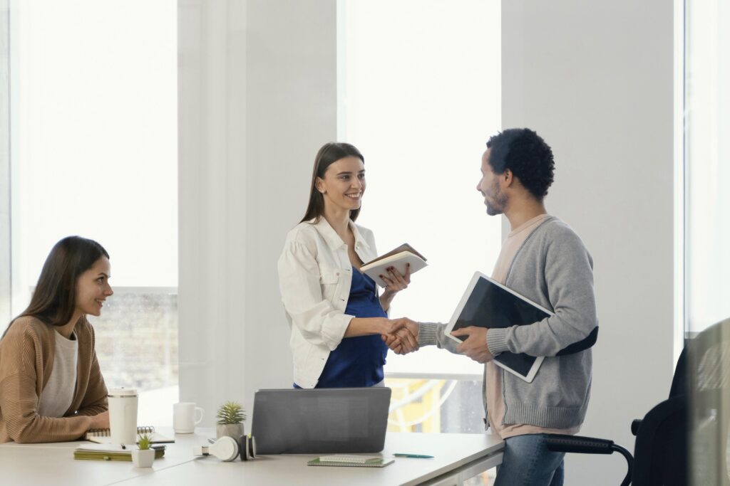 Three people are in a bright office. Two stand, shaking hands and smiling with notebooks and folders—demonstrating how to build a strong candidate pipeline for top administrative talent. One person sits at a laptop, watching them and smiling.