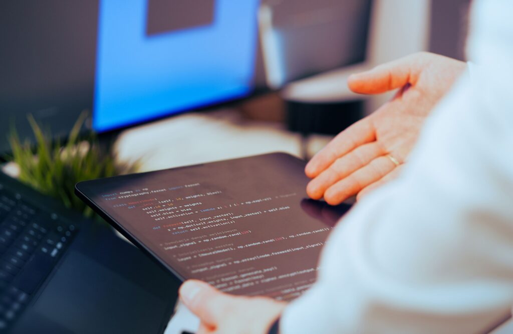 A person gestures towards a tablet displaying lines of computer code, with a laptop and monitor in the blurred background on a desk—illustrating how BCL Search helps AI companies build administrative teams for software development projects.
