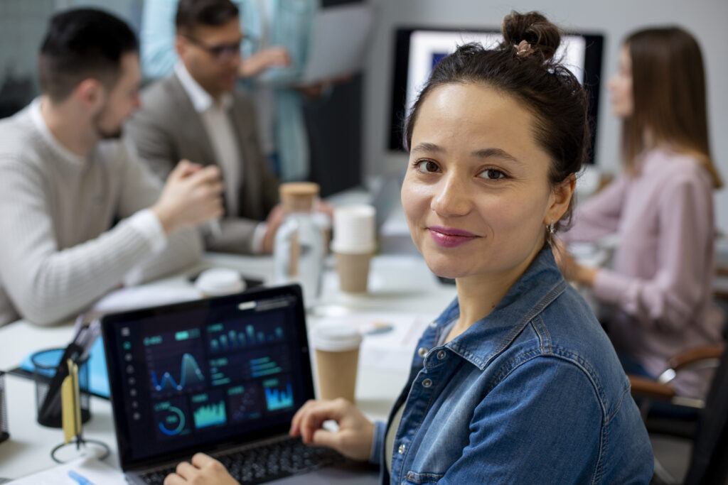 Woman smiles at camera while working on a laptop displaying charts and graphs. She sits at a table with coffee cups and papers, assisting with Executive Assistant Recruiting for Fintech Firms, as colleagues discuss strategies in the background in a modern office setting.